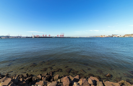 Rocks and water ripples against ships cranes against blue sky at high tide in Durban harbor in South...