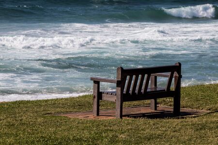 bench on the grass facing the sea with the waves crashing on the beachの写真素材