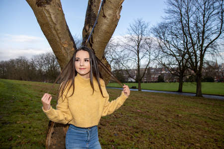 A young attractive woman with long brown hair in the winter time in a wooden park area wearing a yellow jumper looking by a tree in the forestの写真素材