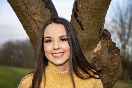 A young attractive woman with long brown hair in the winter time in a wooden park area wearing a yellow jumper looking by a tree in the forestの写真素材