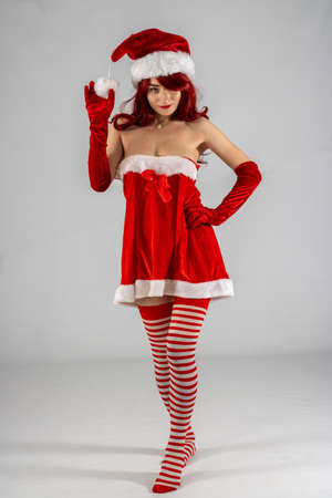 Full length young female attractive model with auburn hair & red lipstick in a studio with a white background wearing a sexy santa outfit with a red Santa hat with red velvet gloves & stripy stockingsの写真素材