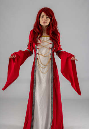 Full length young female attractive model in a studio with a white background, the model is wearing a red medieval costume with red auburn hairの写真素材