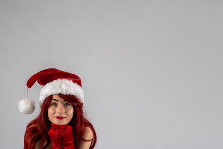 Young female attractive model with auburn hair and red lipstick sat down in a studio with a white background wearing a   santa outfit with a red Santa hat with red velvet gloves.の写真素材