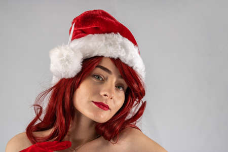 Young female attractive model with auburn hair and red lipstick sat down in a studio with a white background wearing a   santa outfit with a red Santa hat with red velvet gloves.の写真素材