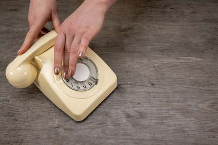 A elegant woman's hand picking up, putting down an old vintage 70's and 80's style rotary telephone receiver on a wooden background, retro office phone call conceptの写真素材