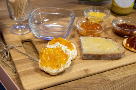 A wooden chopping board on a kitchen work top displaying a selection of jams accompanied with scones, bread and crackersの写真素材