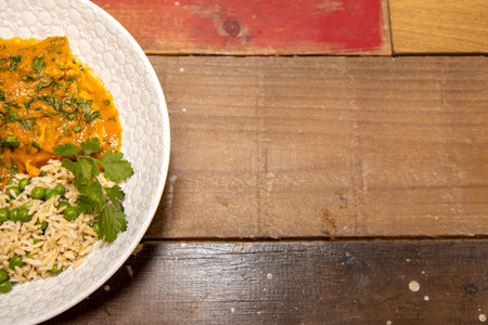 A plate of delicious chicken tikka masala and rice with peas on a wooden kitchen work top backgroundの写真素材