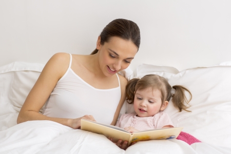 Little girl reading a book with her mother in white bedroomの写真素材