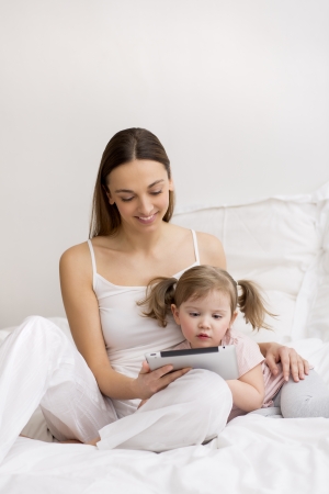 little girl playing digital tablet with her mom in the white bedroomの写真素材