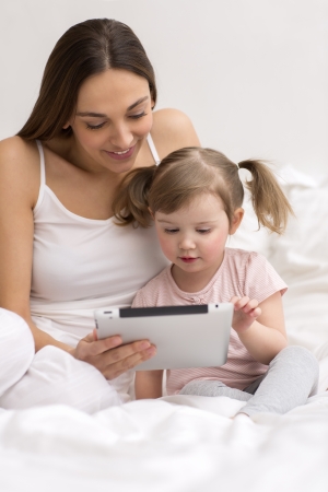 little girl playing tablet with her mom in the white bedroomの写真素材