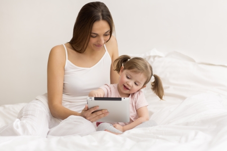 little girl playing tablet with her mom in the white bedroomの写真素材