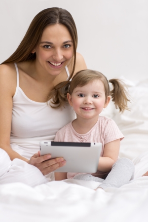 Portrait of a little girl playing tablet with her mom in the white bedroomの写真素材