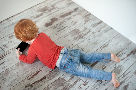 The redheaded child lies on the floor and looks at the cartoon in the phoneの写真素材