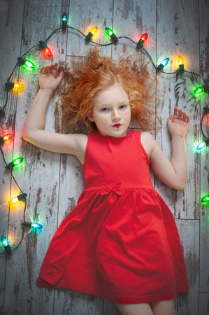 Curly red-haired girl in a red dress lying with a garlandの写真素材