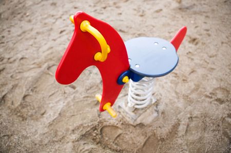 A brightly colored red, blue and yellow rocking horse in a public playground surrounded by sand. - focus on the head with a shallow depth of field.の写真素材