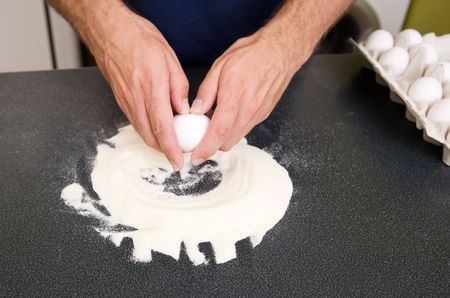 A young man making pasta at home in an apartment kitchen - Cracking an egg into the flour to be...