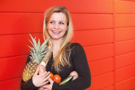 Smiling woman leaning against red wall while holing fruits and vegetablesの写真素材