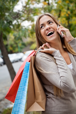 Cheerful young woman communicating on cell phone while carrying shopping bags on shoulderの写真素材