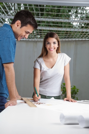 Portrait of young female architect working on blueprint with colleague standing by tableの写真素材