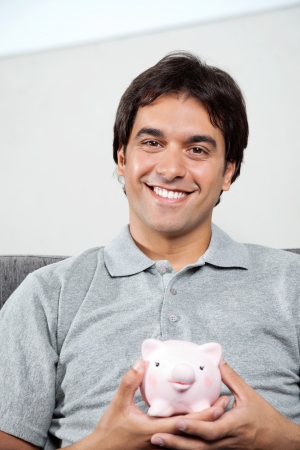 Portrait of young man in casual grey t-shirt smiling while holding piggybankの写真素材