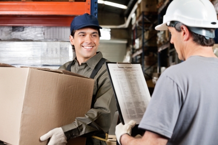 Young warehouse worker with cardboard box looking at male supervisor with clipboardの写真素材