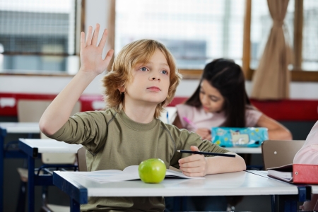 Schoolboy Looking Away While Raising Hand At Deskの写真素材