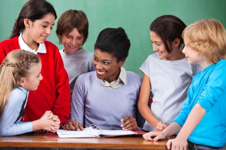 Happy Teacher Teaching Children At Desk In Classroomの写真素材
