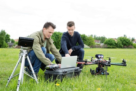 Young male technicians using laptop by tripod and UAV octocopter in park