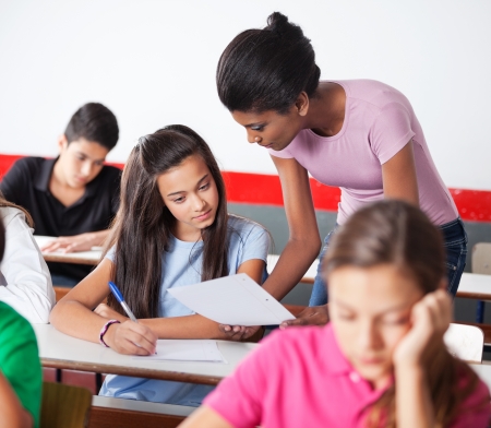 Young female teacher showing paper to teenage girl during examination in classroomの写真素材