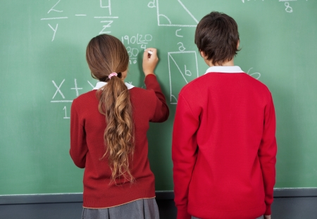 Rear view of teenage girl writing on board with classmate standing in classroomの写真素材