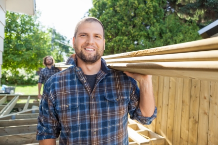Portrait of happy carpenter and coworker carrying lumbers on shoulders at construction siteの写真素材