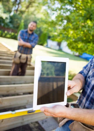 Cropped image of carpenter holding digital tablet with coworker in background at siteの写真素材