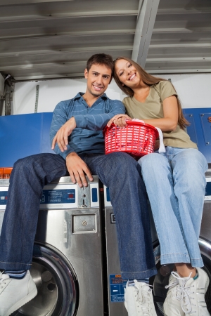 Low angle view of loving young couple with laundry basket sitting on washing machinesの写真素材