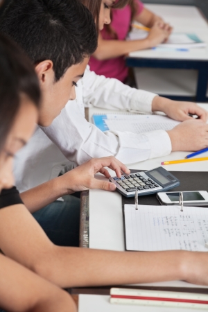 Teenage schoolboy using calculator while studying at desk in classroomの写真素材