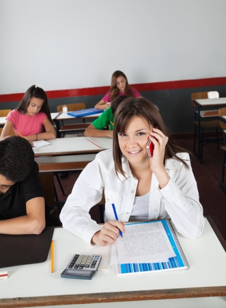 Portrait of teenage schoolgirl using cellphone while sitting at desk in classroomの写真素材