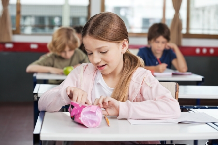 Cute little schoolgirl searching in pouch at desk with classmates の写真素材