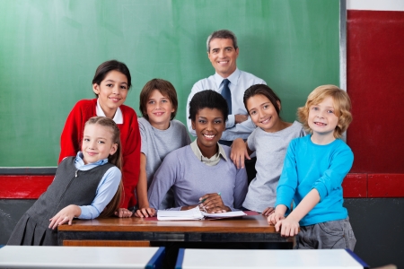 Portrait of happy confident male and female teachers with schoolchildren together at desk in classroomの写真素材