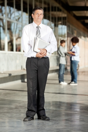 Full length portrait of mature male professor with books standing on college campusの写真素材