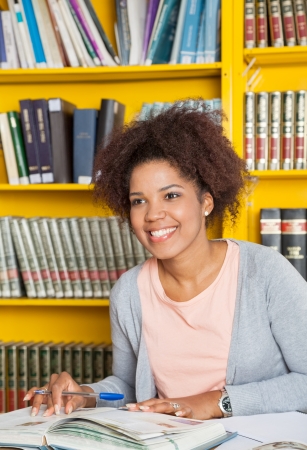 Beautiful college student with books and pen looking away in libraryの写真素材