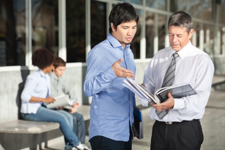 Mature male teacher holding books while discussing with student on college campusの写真素材