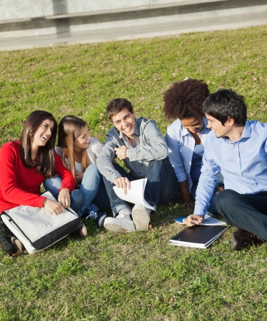 Portrait of happy young man with classmates sitting on grass at college campusの写真素材