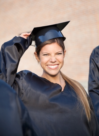 Portrait of cheerful young woman in graduation gown holding mortar board on university campusの写真素材