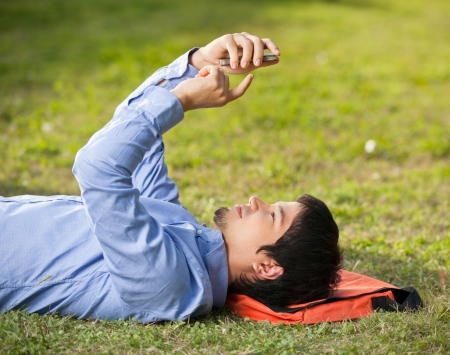 Side view of young man using mobilephone while lying on grass at college campusの写真素材