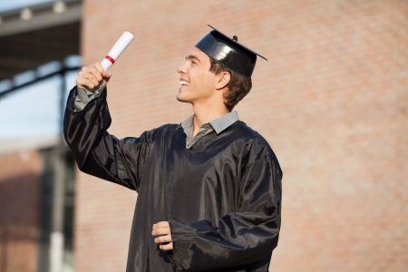 Happy young man in graduation gown looking at certificate on university campusの写真素材