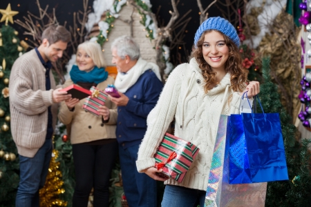 Portrait of beautiful young woman holding Christmas presents and shopping bags with family standing in background at storeの写真素材