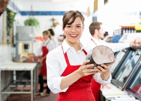 Portrait of happy female butcher holding ham at store with colleagues working in backgroundの写真素材
