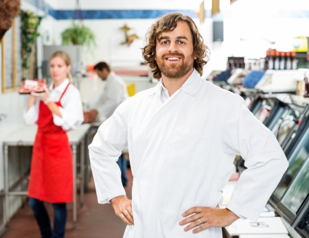 Portrait of confident butcher standing at store with colleagues working in backgroundの写真素材