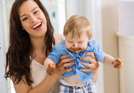 Mid adult mother holding baby boy with cake icing on face and hands at birthday partyの写真素材