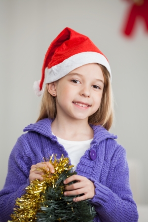 Portrait of smiling girl wearing Santa hat holding tinsels at home during Christmasの写真素材
