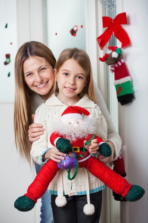 Portrait of loving mother with daughter holding Santa toy while standing by door at homeの写真素材